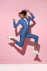 Full body of cheerful young African American female in denim outfit jumping above ground with raised arms and smiling while having fun against pink background during photo session - ADSF43358