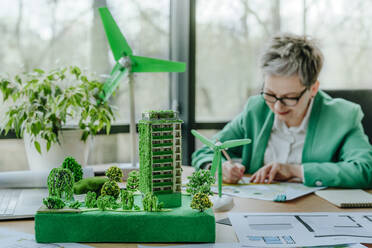 Businesswoman sketching blueprint with biophilic architectural model in office - YTF00609