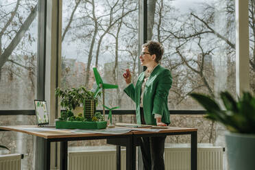 Thoughtful architect standing with biophilic architecture and wind turbine models on table in office - YTF00611