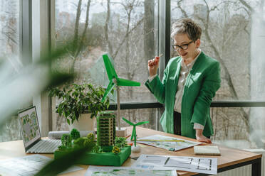 Geschäftsfrau mit Blick auf Blaupausen und biophile Architekturmodelle auf dem Schreibtisch - YTF00612