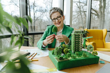 Smiling mature wearing eyeglasses businesswoman holding tree model in office - YTF00616