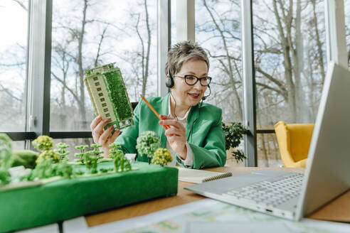 Businesswoman wearing headset showing biophilic architecture model through laptop in office - YTF00617