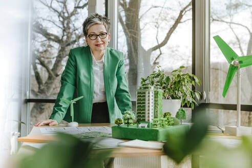 Smiling businesswoman standing with biophilic architectural and wind turbine models on desk at office - YTF00621