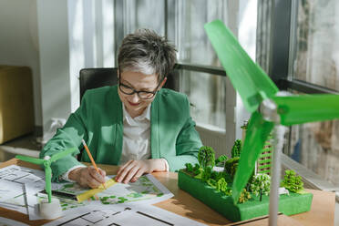 Architect sketching blueprint with wind turbines and biophilic architecture model in office - YTF00622