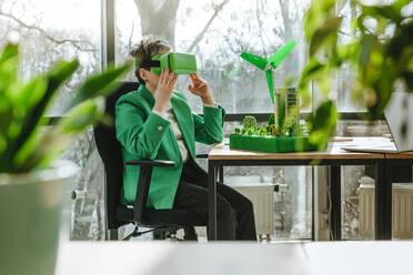 Businesswoman wearing VR glasses sitting with wind turbine and biophilic architectural models in office - YTF00626