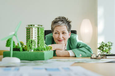 Smiling mature businesswoman looking at biophilic architectural model in office - YTF00635