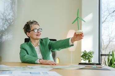 Mature businesswoman holding wind turbine model sitting at desk in office - YTF00639