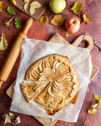 Draufsicht auf einen köstlichen hausgemachten Mürbeteigkuchen auf einem hölzernen Schneidebrett in der Nähe eines Nudelholzes, Herbstblätter und Äpfel - ADSF43372