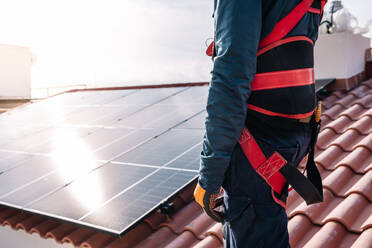 Side view of faceless man in working harness standing on tiled roof with installed solar panel in residential district - ADSF43413