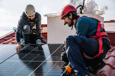 Erwachsene bärtige Arbeiter mit Helmen und Gurten bei der Installation von Sonnenkollektoren auf dem Ziegeldach eines Wohnhauses - ADSF43414