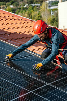 Männlicher Arbeiter mit Schutzhandschuhen und Helm auf dem Dach eines Hauses beim Verschrauben von Solarmodulen während der Installation auf dem Dach - ADSF43415