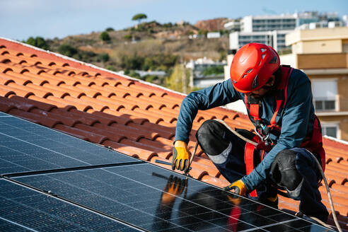 Männlicher Arbeiter mit Schutzhandschuhen und Helm auf dem Dach eines Hauses beim Verschrauben von Solarmodulen während der Installation auf dem Dach - ADSF43417