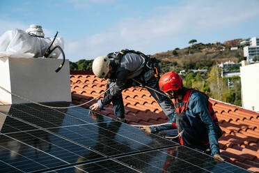 Erwachsene bärtige Arbeiter mit Helmen und Gurten bei der Installation von Sonnenkollektoren auf dem Ziegeldach eines Wohnhauses - ADSF43419