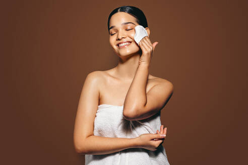 Junge Frau mit klarer, strahlender Haut, die ihr Gesicht mit einem Schwamm reinigt; Peeling als Teil ihrer Schönheitsroutine. Frau in einem Badetuch, die lächelt und ihre Hautpflegeroutine genießt. - JLPSF29372