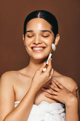 Lächelnde Frau, die in einem Studio ihr makelloses Gesicht mit einem Jadewalzen massiert. Eine Frau benutzt einen Schönheitsstein, um die Durchblutung ihrer Haut anzuregen und ein perfektes Strahlen zu erzielen. Hautpflegeroutine für gesunde Haut. - JLPSF29377
