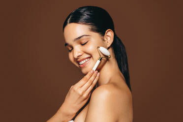 Young woman massaging her skin with a jade stone roller in a studio, she smiles as she uses this beauty tool. This woman is enjoying pampering her skin, and she feels confident in her routine. - JLPSF29378