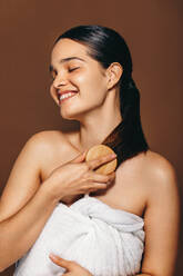 Young woman smiles as she brushes her hair after a bath, she is wearing a bath towel in a studio. This beautiful woman knows the importance of self-care and grooming, and she takes pride in her beauty routine. - JLPSF29379