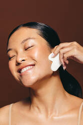 Happy young Asian woman smiles as she massages her face using a gua sha beauty tool. Using this tool to keep her skin youthful, she radiates confidence and self-assurance. - JLPSF29406