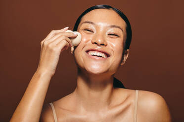 Woman with radiant skin smiling as she applies face oil with a beauty sponge. Happy young woman gently massaging and blending the moisturizing treatment into her skin in the midst of her skincare routine. - JLPSF29409