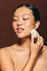 Female with glowing skin applies a facial treatment using a beauty sponge, she gently massages her skin with the blender. With a self-assured expression on her face, she is confident in her skincare routine. - JLPSF29410