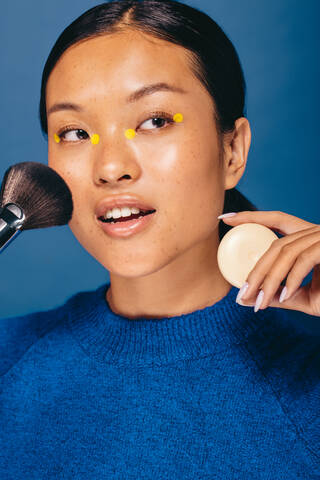 Confident young woman holding a makeup brush and foundation in a studio,  she is wearing four
