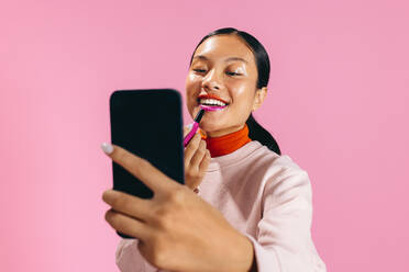 Female using a smartphone as a mirror to apply lipstick in a studio. Gen Z woman doing the two toned lip trend to create a bold and vibrant makeup look on her face. - JLPSF29459