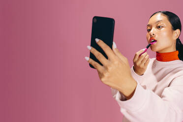 Modische Frau, die ein Smartphone als Spiegel benutzt, um während ihrer Make-up-Routine Lippenstift aufzutragen. Junge Frau in den 20ern, die einen gewagten, zweifarbigen Lippen-Look kreiert, ein modischer Make-up-Trend. - JLPSF29461