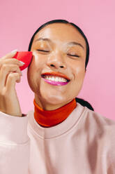 Young woman smiles happily as she blends makeup on her face using a beauty sponge. Woman in her 20’s enjoying her beauty routine, she massages the soft blender on her skin to apply the cosmetic product. - JLPSF29463