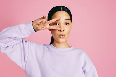 Young woman looking at the camera while doing a peace sign and puckering her lips, making a funny expression. This carefree woman is enjoying herself and flaunting her self confidence and positivity. - JLPSF29476