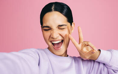 A joyful young woman captures her self-confidence and positivity by taking a selfie in a studio, flashing a peace sign - JLPSF29480