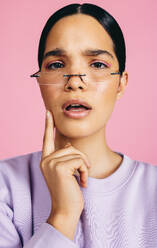 Young woman in her 20s looking at the camera with an inquisitive expression, she is wearing eyeglasses and they are perched on her nose. Thoughtful woman with makeup on her face stands in a studio. - JLPSF29483