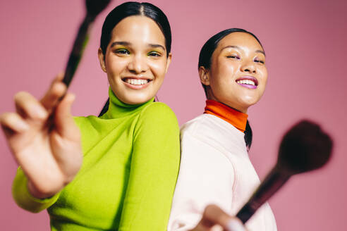 Zwei junge Frauen lächeln in die Kamera, während sie ihre Schönheitspinsel in der Hand halten und ein leuchtendes Make-up tragen. Glamouröse Frauen, die Spaß haben und ihre Liebe für Schönheit und Kosmetik feiern. - JLPSF29494