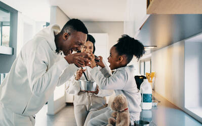 Vater und Tochter spielen zusammen ein Spiel, während Mutter in der Küche das Frühstück zubereitet. Vater und Tochter haben ein tolles Verhältnis zueinander und Mutter lächelt. Glückliche schwarze Familie, die Zeit miteinander zu Hause verbringt. - JLPSF29504