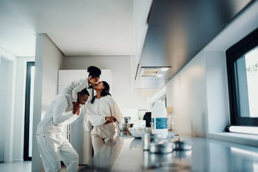 Family fun at breakfast time. Mom kisses her daughter from dad’s shoulders. Family having playful moments as mom prepares breakfast in the morning. - JLPSF29507