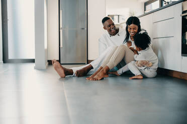 Eine dreiköpfige Familie, die zusammen spielt und einen lustigen Moment genießt. Eltern, die mit ihrer Tochter Spaß haben, während sie in der Küche auf dem Boden sitzen. Ein schwarzes Paar, das mit seinem Kind zu Hause zusammen ist. - JLPSF29511