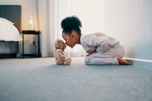 Ein süßes kleines Mädchen, das seine Kindheit zu Hause mit seinem Lieblingsspielzeug, einem niedlichen Teddybär, genießt. Sie berührt die Nasen des Stofftiers und genießt ihre Freizeit und die Unschuld der Kindheit. - JLPSF29527