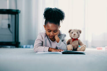 Elementary age girl lies on the floor in her bedroom at home, she is reading a book and has her teddy bear next to her. She is focused on learning and education during this time of homeschooling. - JLPSF29528