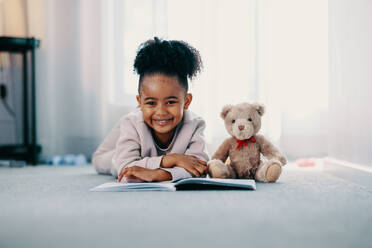 Ein junges Mädchen mit einem süßen Lächeln im Gesicht blickt in die Kamera, während sie zu Hause neben ihrem Teddybär auf dem Boden liegt und ihre Freizeit genießt, während sie durch Lesen lernt und wächst. - JLPSF29529