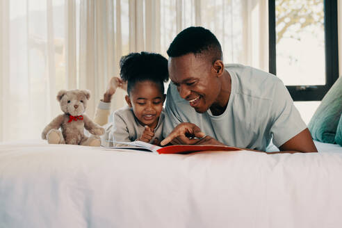 Papa liest seiner Tochter am Wochenende eine Geschichte vor. Vater und Tochter liegen mit einem Buch auf dem Bett. Ein Elternteil verbringt viel Zeit mit seinem Kind und fördert seine Lesekompetenz. - JLPSF29530