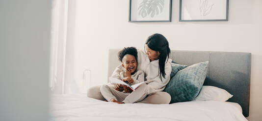 Mom and daughter laughing together as they read a story book on a bed. Young girl in elementary age is getting entertained by a fairytale she’s reading with her mother. - JLPSF29534