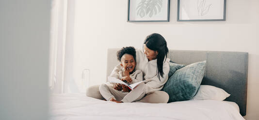 Mutter und Tochter lachen zusammen, während sie auf einem Bett ein Märchenbuch lesen. Ein junges Mädchen im Grundschulalter amüsiert sich über ein Märchen, das sie mit ihrer Mutter liest. - JLPSF29534