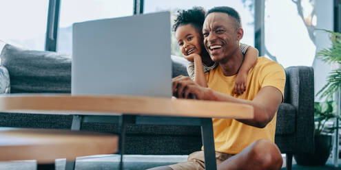 Vater und Tochter lächeln, während sie sich ein interessantes Video auf einem Online-Kinderkanal ansehen. Vater und Tochter surfen gemeinsam zu Hause im Internet. Familienunterhaltung mit moderner Technologie. - JLPSF29541