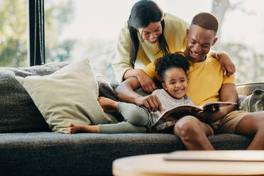 Glückliches kleines Mädchen, das mit seiner Mutter und seinem Vater eine Geschichte liest. Eltern, die ihrem Kind zu Hause Aufmerksamkeit schenken. Lustige Familienmomente am Wochenende. - JLPSF29545