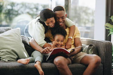 Parents reading a book with their daughter on a couch. Happy family spending time together at home. African couple raising a toddler in a loving environment. - JLPSF29546
