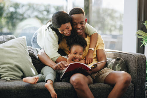 Eltern, die mit ihrer Tochter auf einer Couch ein Buch lesen. Glückliche Familie, die zu Hause Zeit miteinander verbringt. Afrikanisches Paar, das ein Kleinkind in einer liebevollen Umgebung großzieht. - JLPSF29546