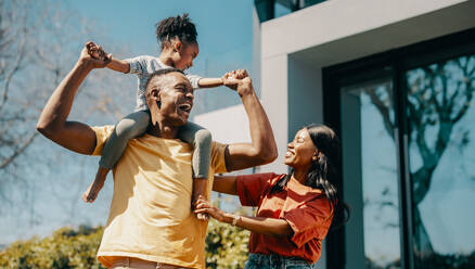 Family fun time with mom and dad at home. Couple playing with their daughter and creating happy childhood memories. Dad carrying his daughter on his shoulders and standing next to mom. - JLPSF29548