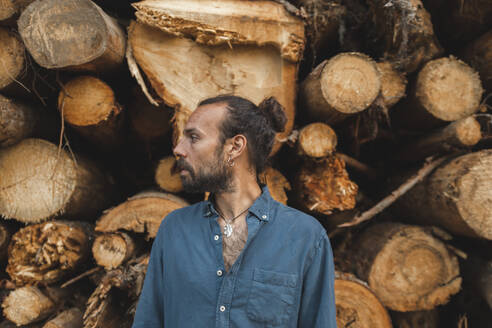 Nachdenklicher Mann mit Haarknoten vor Holzstämmen im Sägewerk - PCLF00257