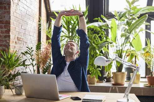 Relaxed businessman stretching arms after work at loft office - BSZF02289