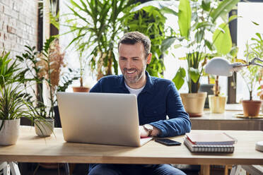 Happy businessman working on laptop at loft office - BSZF02291