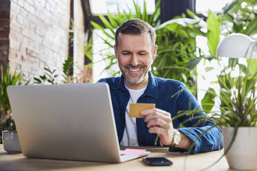 Lächelnder Geschäftsmann mit Laptop und Kreditkarte im Loft-Büro - BSZF02295